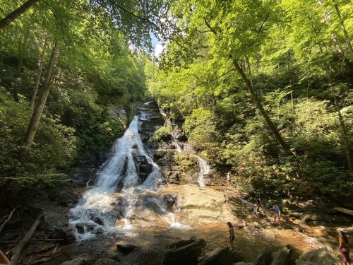 A serene waterfall cascades down rocky terrain, surrounded by lush green trees and people enjoying the water below.