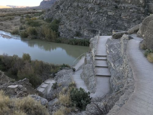 A winding pathway leads down rocky terrain to a calm river, surrounded by lush greenery and hills in the background.