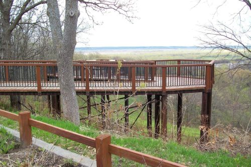 A wooden observation deck on stilts overlooking a green landscape and water in the distance, surrounded by trees.