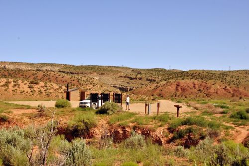 A person stands near a parking area surrounded by red rock formations and sparse vegetation under a clear blue sky.