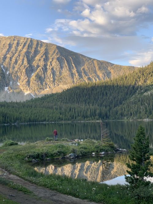 A person stands by a serene lake, surrounded by mountains and lush greenery, reflecting the landscape in the water.