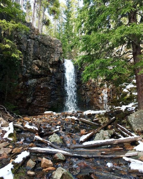 A serene waterfall cascades over rocks, surrounded by lush greenery and patches of snow in a tranquil forest setting.