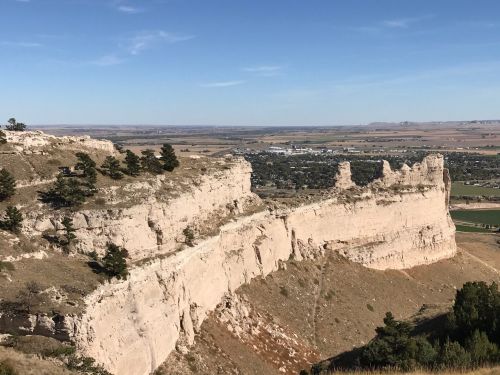 A rocky cliff with trees on top overlooks a valley and distant farmland under a clear blue sky.