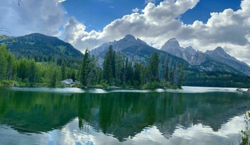 A serene lake reflects towering mountains and lush trees under a partly cloudy sky.