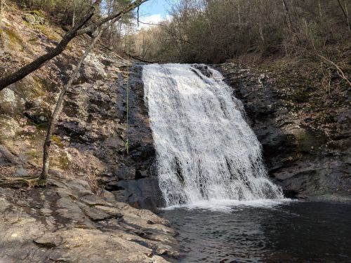 A serene waterfall cascading over rocky terrain, surrounded by trees and a clear blue sky.