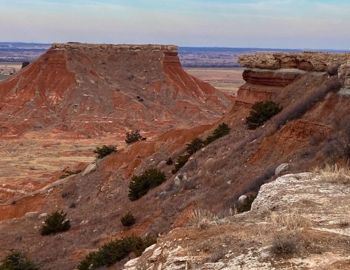 A rugged landscape featuring red rock formations and cliffs, with sparse vegetation and a distant horizon under a cloudy sky.