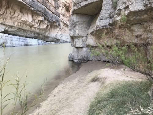 A serene riverbank scene with rocky cliffs and gentle water, surrounded by sparse vegetation.