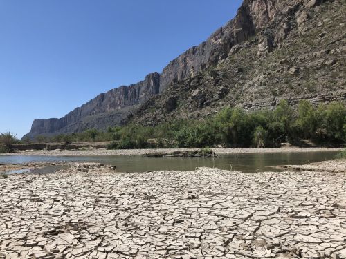 A dry, cracked landscape with a river and steep rocky cliffs under a clear blue sky.