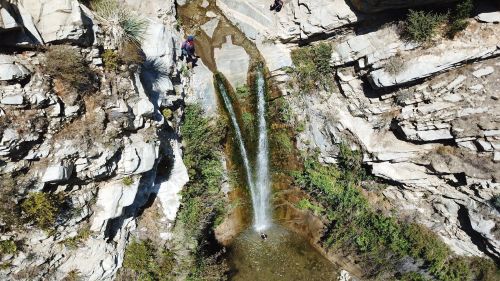 Aerial view of a waterfall cascading into a rocky pool, surrounded by steep cliffs and lush greenery.
