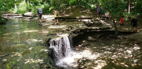A small waterfall flows over rocks in a wooded area, with hikers exploring the stream and surrounding greenery.