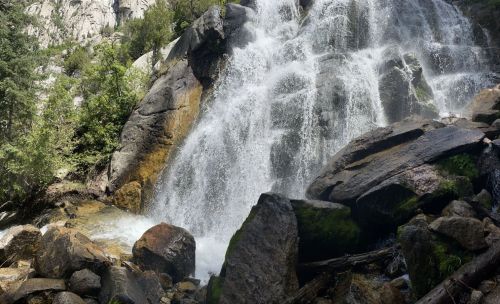 A cascading waterfall flows over large rocks, surrounded by lush greenery and towering cliffs.