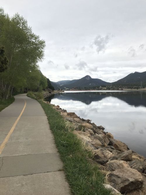 A serene lakeside path lined with trees, reflecting mountains and clouds in the calm water.