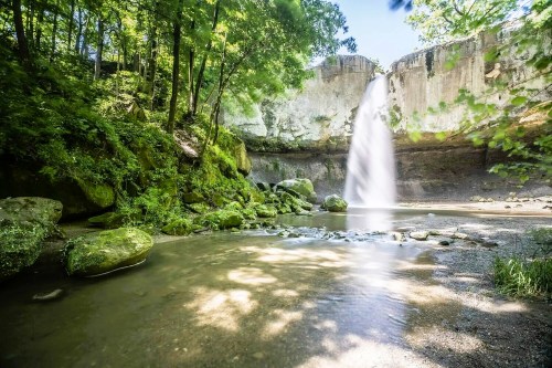 A serene waterfall cascades over a rocky cliff, surrounded by lush greenery and a calm river below.