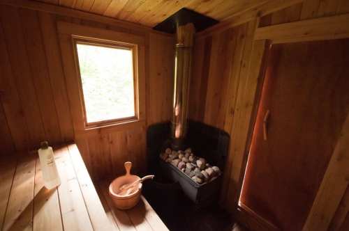 Interior of a wooden sauna featuring a stone stove, a window, and a wooden door. A bucket and water bottle are visible.