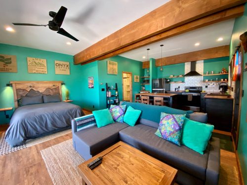 Cozy room featuring a bed, sectional sofa with colorful pillows, and a modern kitchen in a vibrant teal setting.