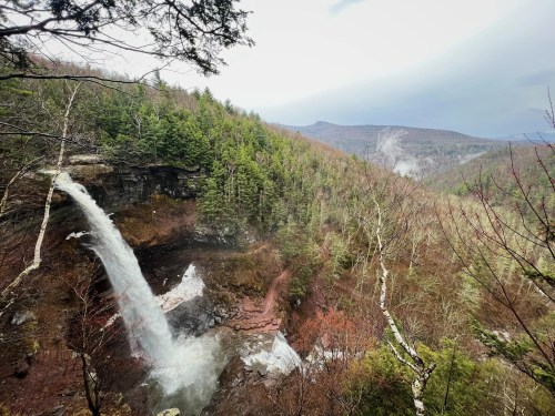 A scenic view of a waterfall cascading down rocky cliffs, surrounded by lush green trees and misty mountains in the background.