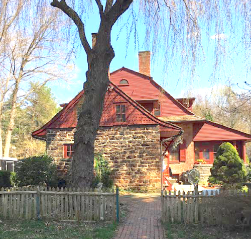 A charming two-story house with a stone base and red wooden upper, surrounded by greenery and a picket fence.