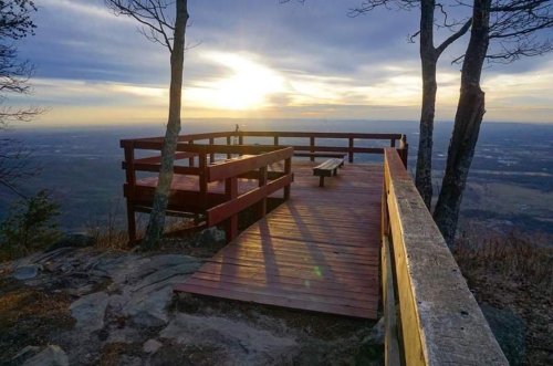 A wooden observation deck with benches overlooks a valley at sunset, framed by trees and a cloudy sky.