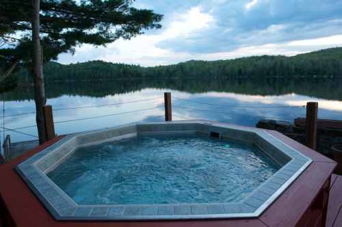 A serene hot tub overlooking a calm lake surrounded by lush greenery and a cloudy sky.