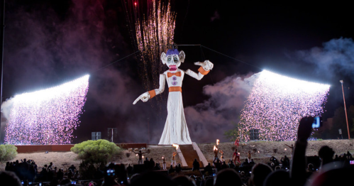 A large puppet figure with fireworks and colorful lights, surrounded by an audience at a nighttime event.