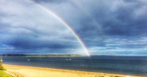 A vibrant rainbow arcs over a calm sea, with a sandy beach and cloudy sky in the background.