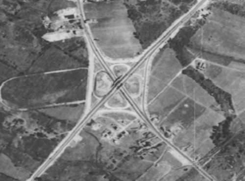 Aerial view of a highway interchange surrounded by fields and scattered structures.
