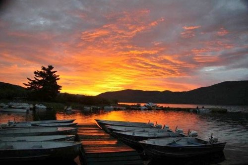 A serene lake at sunset, with colorful clouds reflecting on the water and boats lined up on the shore.