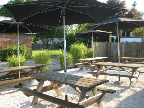 Outdoor seating area with wooden picnic tables and large black umbrellas, surrounded by greenery and gravel.
