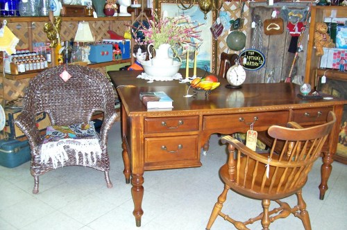 A vintage wooden desk with two chairs, surrounded by various antique items and decor in a cozy shop setting.