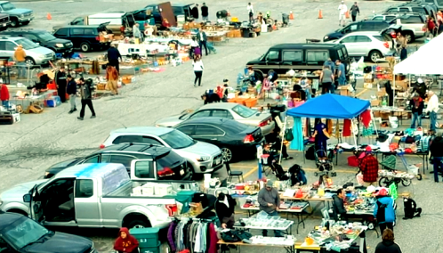A bustling flea market with various stalls, cars parked nearby, and people browsing through items for sale.
