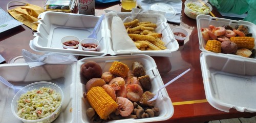 A table with takeout containers filled with shrimp, corn, potatoes, coleslaw, and fries, accompanied by dipping sauces.