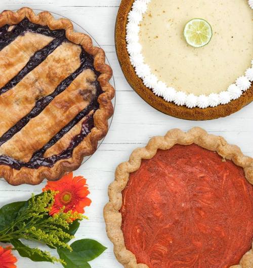 Three delicious pies: a blueberry pie, a lime pie, and a pumpkin pie, arranged with fresh flowers on a wooden surface.