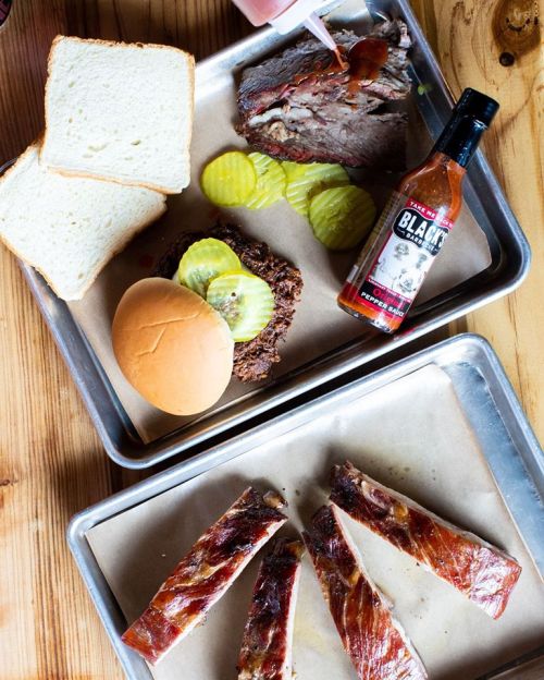 A wooden table with trays of barbecue brisket, ribs, pickles, bread, and hot sauce.