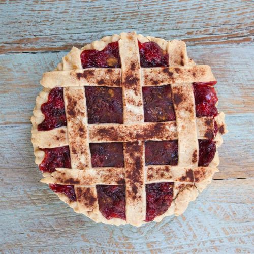 A freshly baked pie with a lattice crust, filled with dark red fruit filling, on a rustic wooden surface.