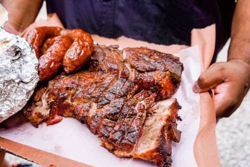 A person holds a tray with smoked ribs, sausage, and a foil-wrapped item, showcasing delicious barbecue food.