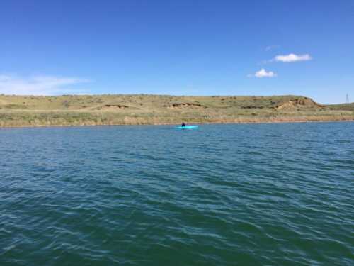 A calm lake with a person kayaking in the distance, surrounded by green hills under a clear blue sky.