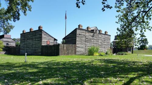 Historic log cabins with a flag in the background, surrounded by green grass and scattered leaves under a clear blue sky.