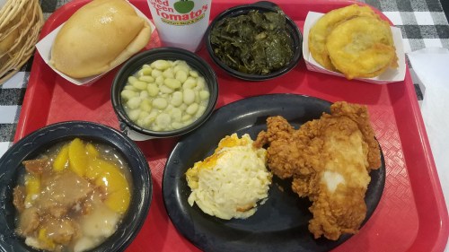 A red tray with fried chicken, macaroni and cheese, collard greens, lima beans, peach cobbler, a roll, and a drink.