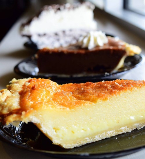 Three slices of pie on black plates: a golden custard pie, a chocolate pie, and a cream pie in the background.