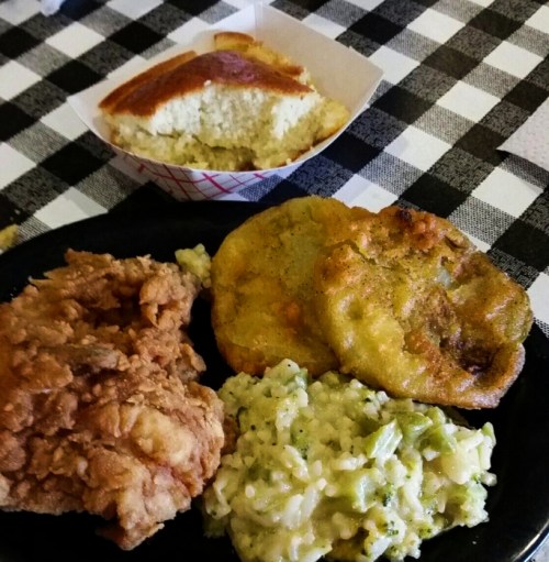 A plate of fried chicken, rice with broccoli, fried plantains, and a side of cornbread on a checkered tablecloth.