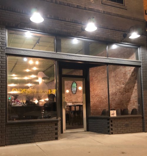 Exterior view of a restaurant with large windows, warm lighting, and a brick facade, showcasing a welcoming atmosphere.