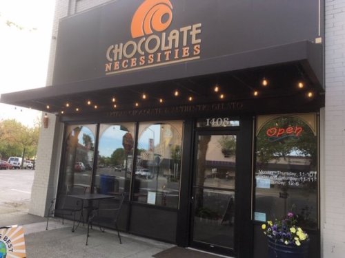 Exterior of "Chocolate Necessities" shop with a sign, open hours, and outdoor seating area.