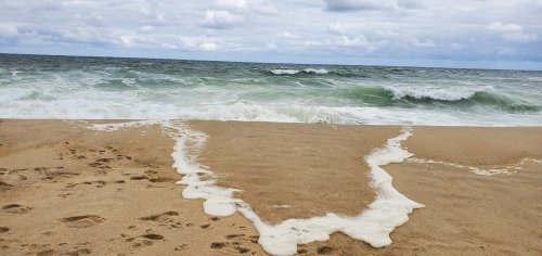 Waves gently wash onto a sandy beach, leaving foam along the shore under a cloudy sky.