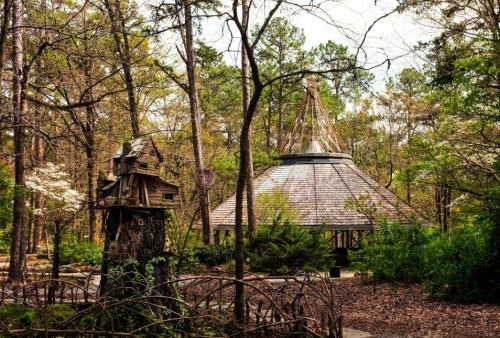 A whimsical treehouse beside a circular pavilion, surrounded by lush trees and greenery in a serene forest setting.