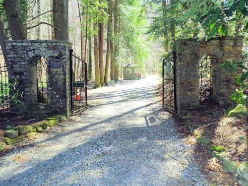 A stone gate opens to a gravel path lined with trees, leading to a cabin in a wooded area.