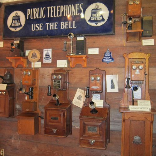 A display of vintage public telephones mounted on a wooden wall, featuring various styles and a "Bell System" sign.