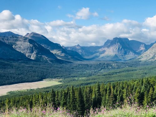 A panoramic view of mountains and valleys, with lush green forests and a blue sky dotted with clouds.