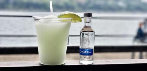 A refreshing cocktail in a plastic cup with lime slices, next to a bottle of Camarena Silver tequila by a waterfront.