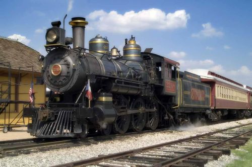 A vintage steam locomotive with a passenger car, parked on tracks under a blue sky with fluffy clouds.