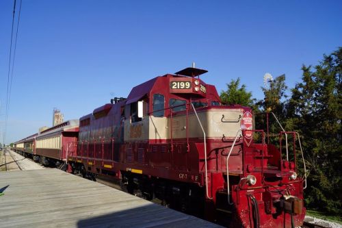 A red and silver locomotive parked beside a wooden platform, with trees and a clear blue sky in the background.